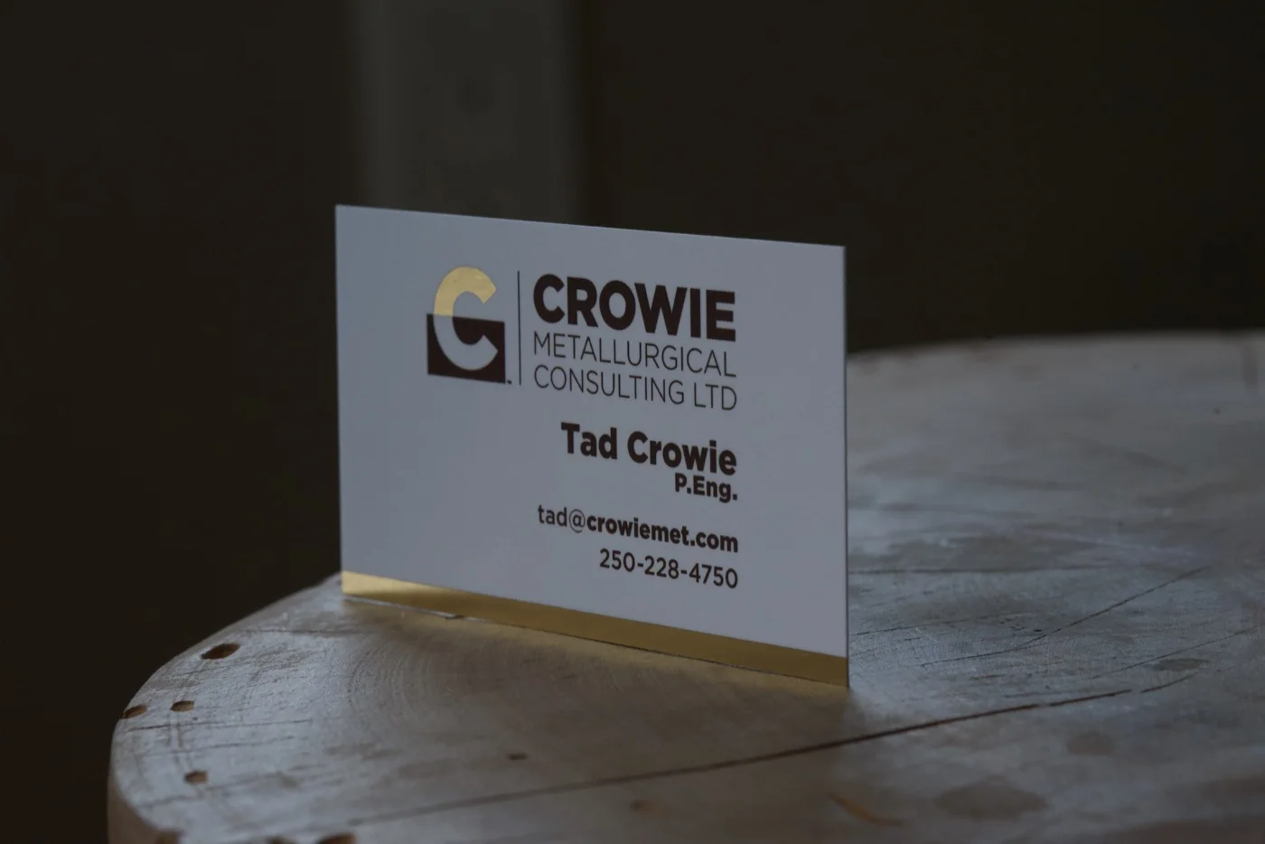 A close-up photo of a printed Crowie Metallurgical Consulting business card resting on a wood round, showing the gold-foil C catching the light, the brown block and wordmark crisply printed on the white card stock, and the gold-edged side of the card visible at the bottom