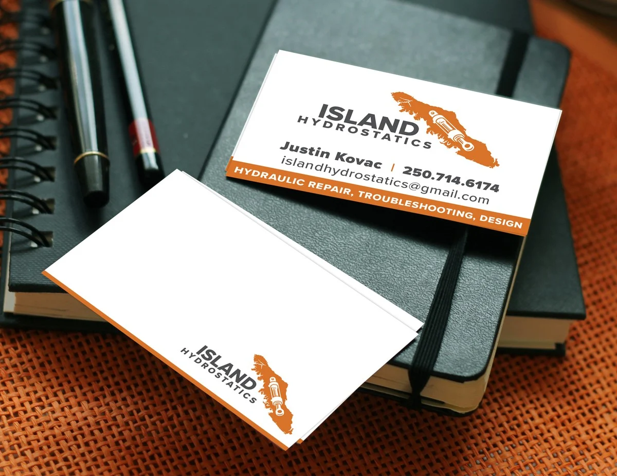 A pair of Island Hydrostatics business cards arranged on a black leather Moleskine notebook with a fountain pen, the front card showing the Vancouver Island silhouette mark above Justin Kovac's contact details and a service strip reading HYDRAULIC REPAIR TROUBLESHOOTING DESIGN, the back card showing the smaller mark on a clean white field